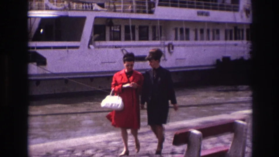 A closer 1967 France frame shows two women conversing as they pass the bench, with the docked white riverboat behind them.