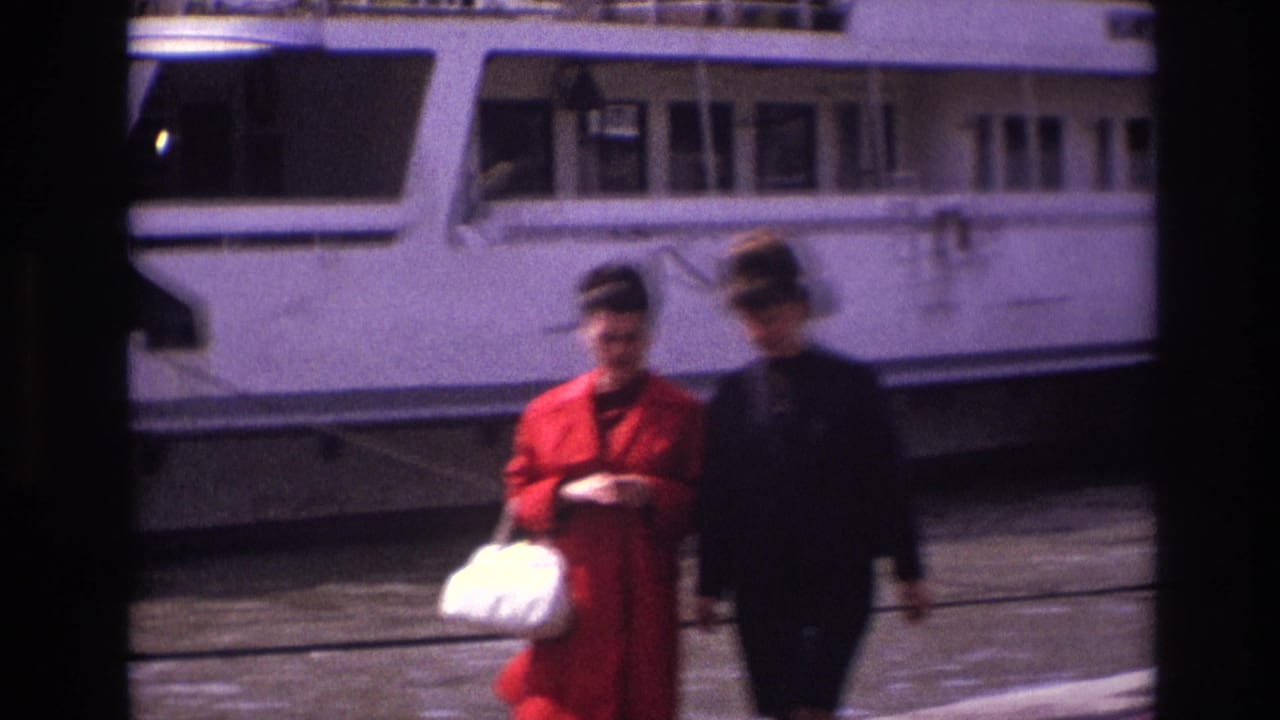 Two adult women in 1967 France walk along a waterside pier, one in a red coat carrying a white handbag, with a large white riverboat behind them.