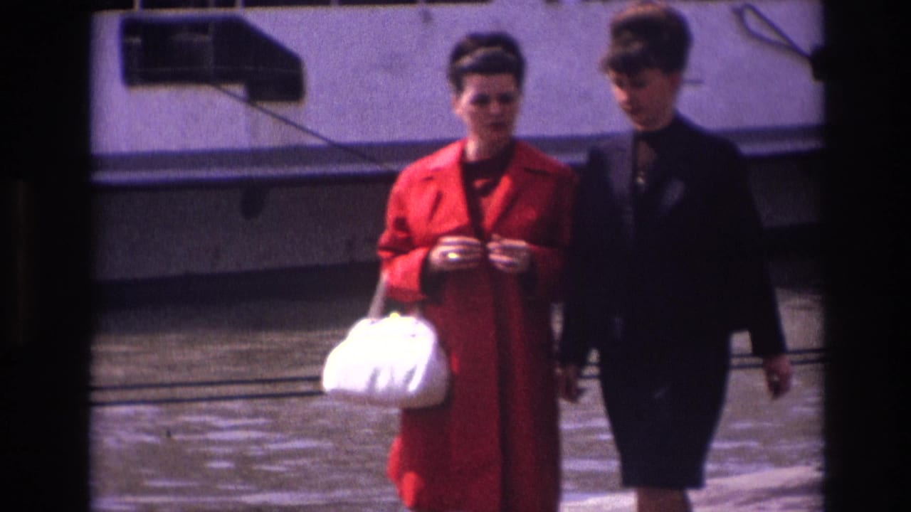 Two adult women in 1967 France pass along the pier, with a red coat, white handbag, dark tailored clothing, and a white riverboat visible behind them.