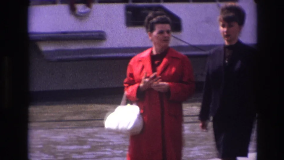 Two adult women in 1967 France walk side by side near the water, one holding a white handbag while the other wears a dark outfit beside a boat hull.