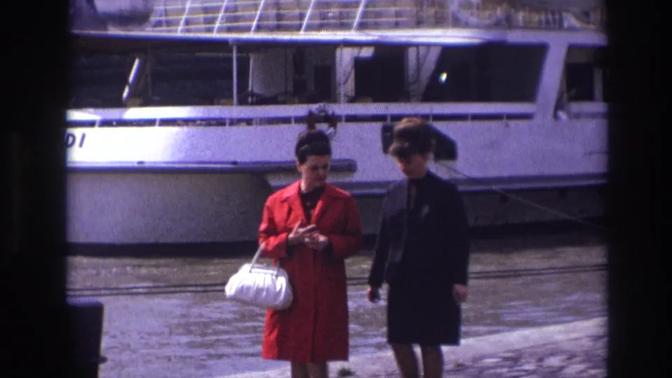 Two adult women in 1967 France continue walking along the pier, framed lower in the shot with the white riverboat, water, and partial lettering behind them.