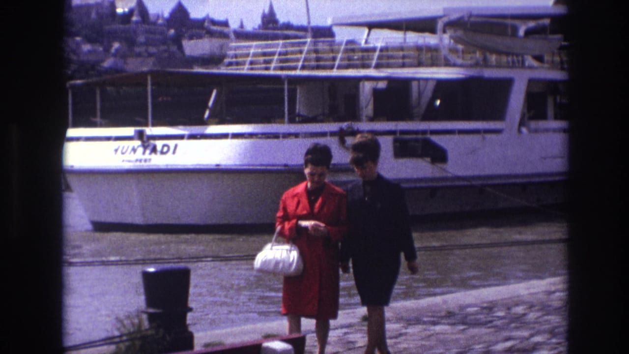 1967 France home-movie frame of two young women walking near a docked white river boat marked HUNYADI, with water and spires behind them.