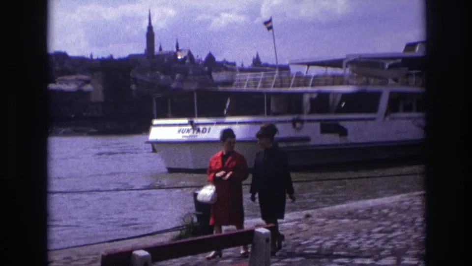 1967 France riverside frame showing two women walking past the HUNYADI boat, with mooring lines, water, and a spired skyline in the background.