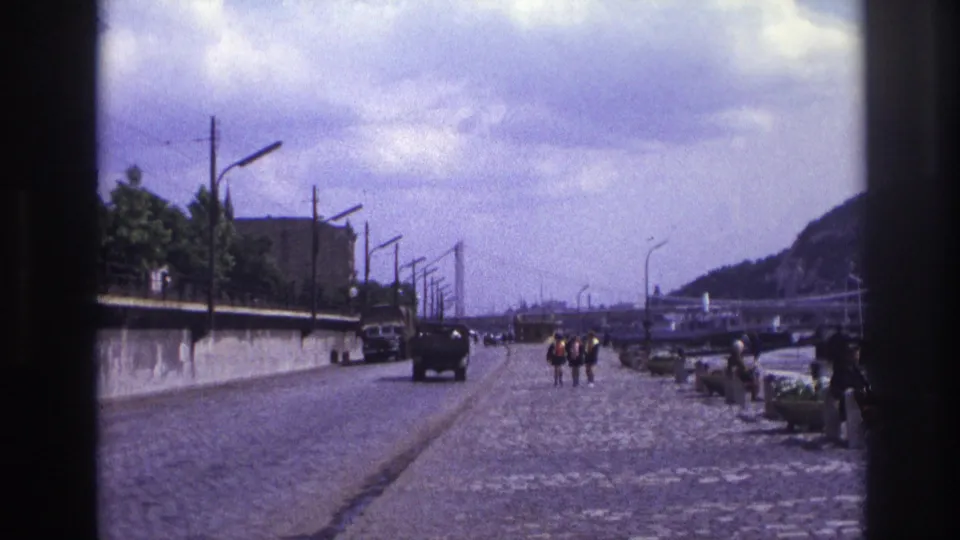Wide 1967 France home-movie view of a riverside road and promenade with older cars, pedestrians, streetlamps, water, hills, and distant bridges or structures.