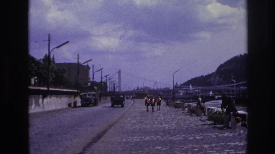 1967 France frame looking down a waterfront promenade, with pedestrians in the distance, older cars, river edge, streetlamps, hills, and overcast sky.