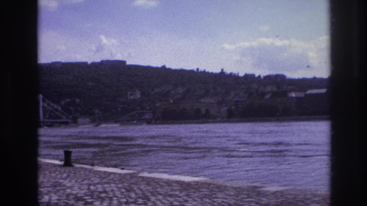 1967 France 8mm frame showing a broad river, cobblestone riverbank, distant hillside, low town buildings, and clouds.