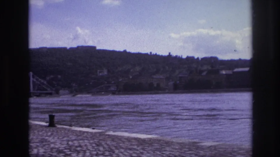 1967 France 8mm frame showing a broad river, cobblestone riverbank, distant hillside, low town buildings, and clouds.