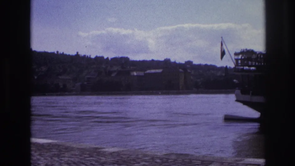 1967 France frame showing a docked passenger boat with a flag beside the river and hillside buildings beyond.