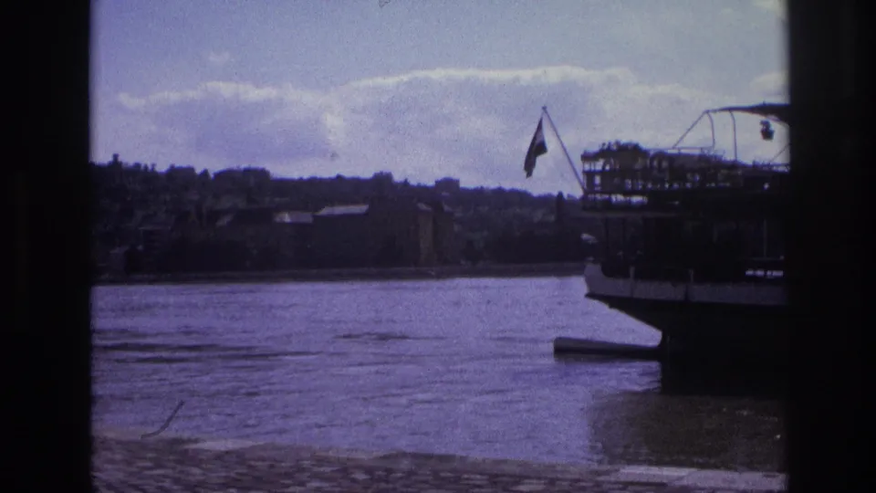 1967 France 8mm frame of a docked passenger boat at the river edge with a flag and hillside town behind it.