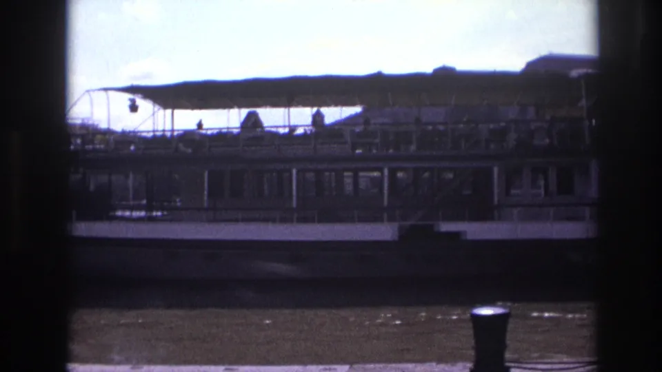 1967 France 8mm view of the side of a docked passenger boat, with windows, canopy, deck railings, river water, and quay edge.