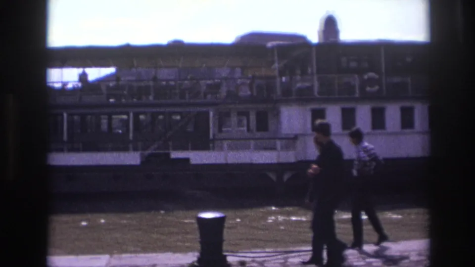 1967 France frame showing pedestrians on a riverside quay with a bollard, water, and a docked passenger boat behind them.