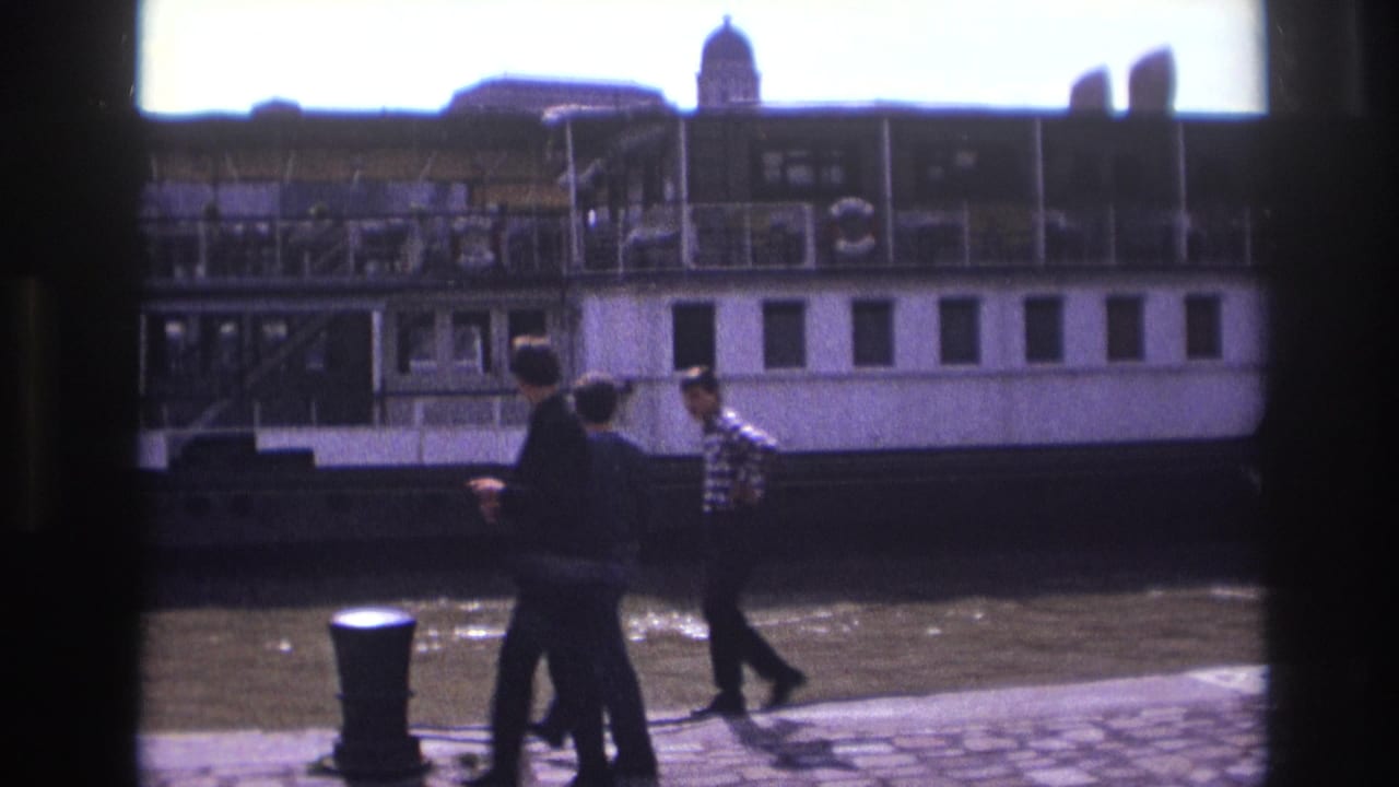 In 1967 France, three men walk along a cobblestone riverside quay with a large white paddlewheel boat in the background.