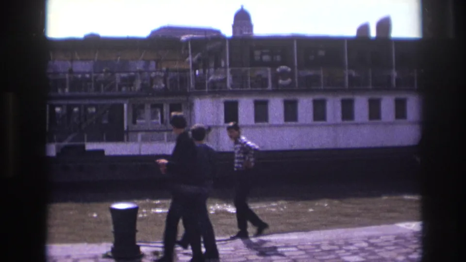 In 1967 France, three men walk along a cobblestone riverside quay with a large white paddlewheel boat in the background.
