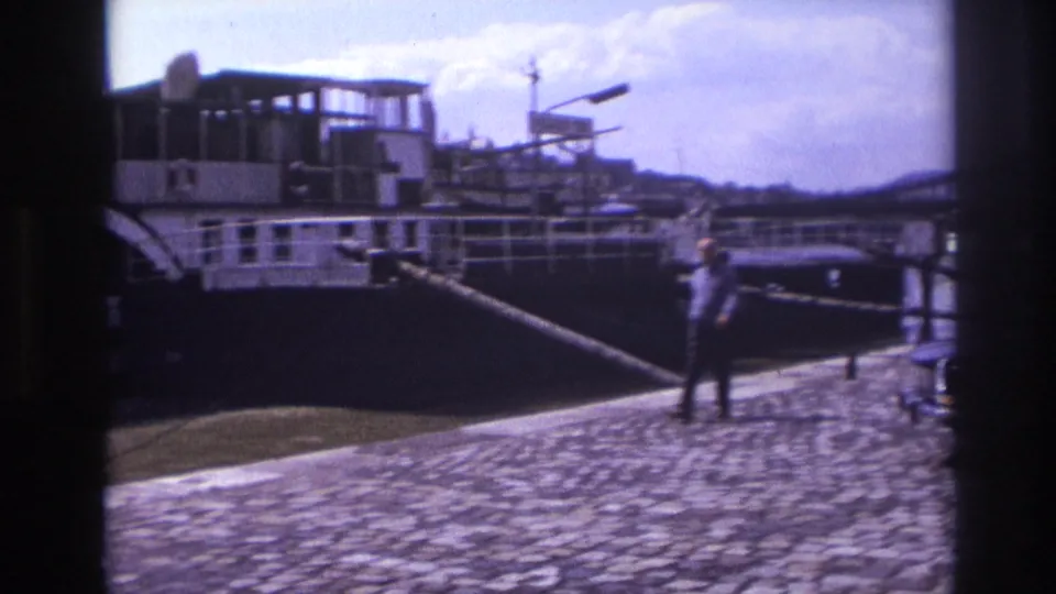The camera looks down the cobblestone quay toward the moored paddlewheel boat, railings, water, and two men near the edge.