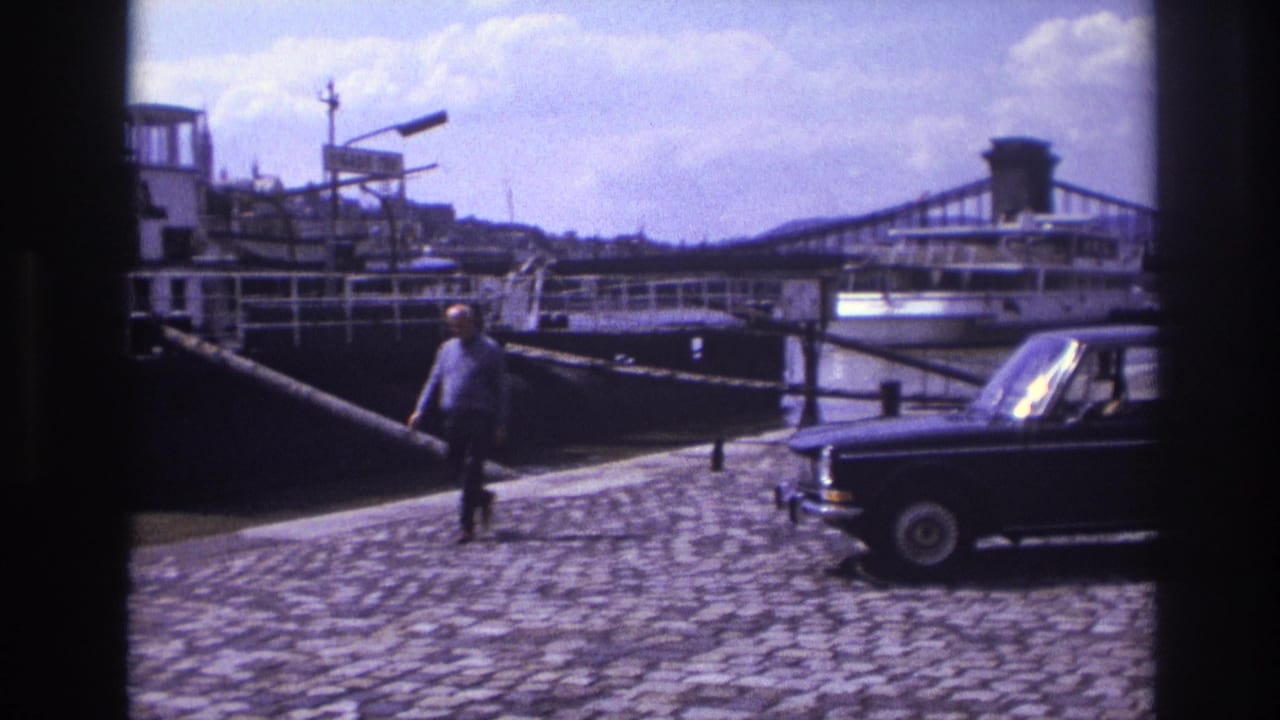1967 France home-movie frame showing a dark sedan on a cobblestone quay, a man walking, a moored riverboat, and a bridge beyond.