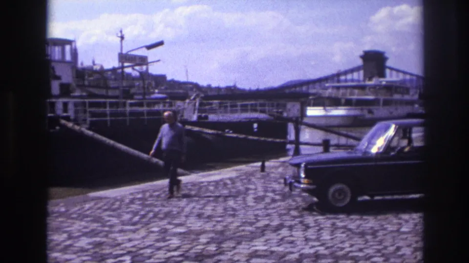 1967 France home-movie frame showing a dark sedan on a cobblestone quay, a man walking, a moored riverboat, and a bridge beyond.
