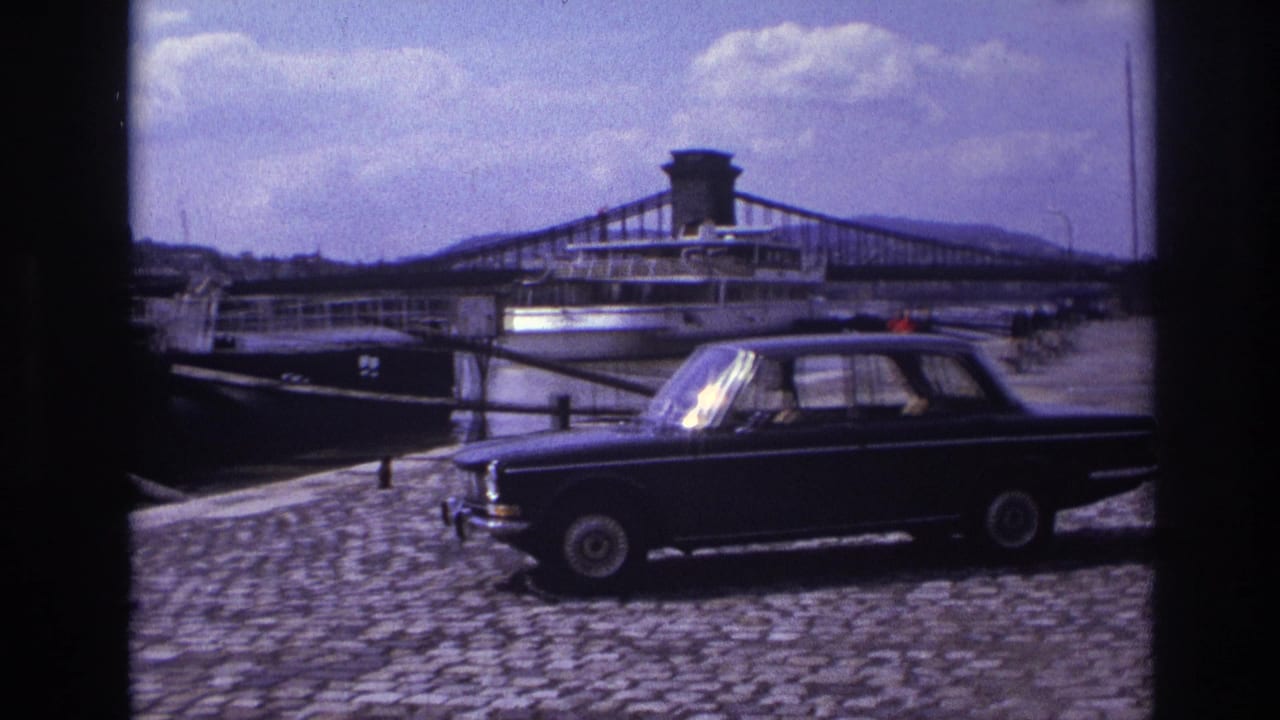 Parked dark sedan beside a riverside pier, with a docked boat and bridge in the background.