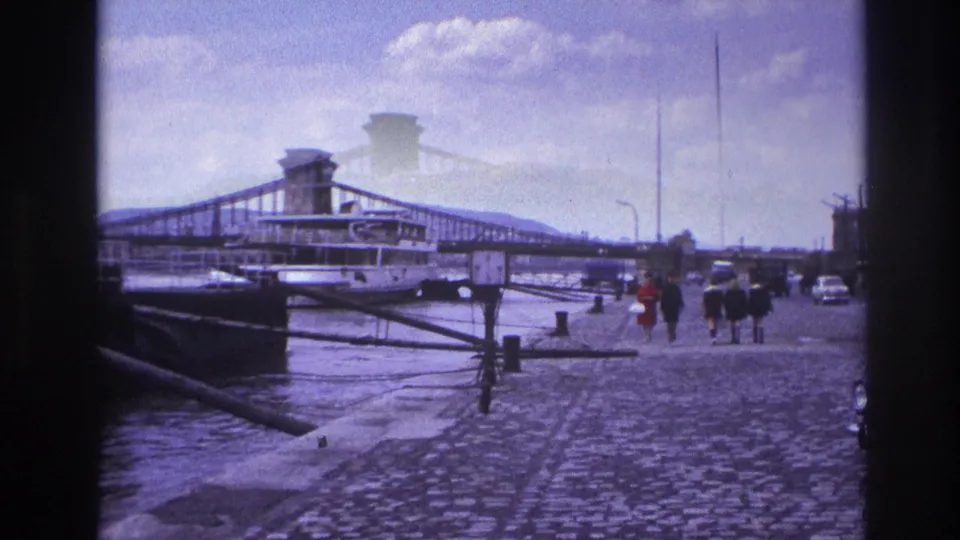 1967 France home-movie view of pedestrians on a cobblestone riverside promenade near a moored boat and bridge.