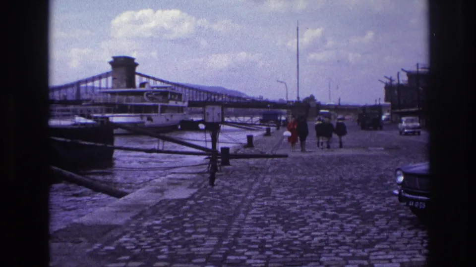 1967 France waterfront frame with pedestrians on a cobbled quay, a bridge over the river, a moored boat, and cars at the edge.