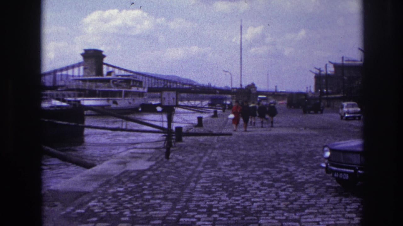 1967 France home-movie frame showing pedestrians on a cobblestone quay beside water, with a suspension bridge, riverboat, and cars visible.