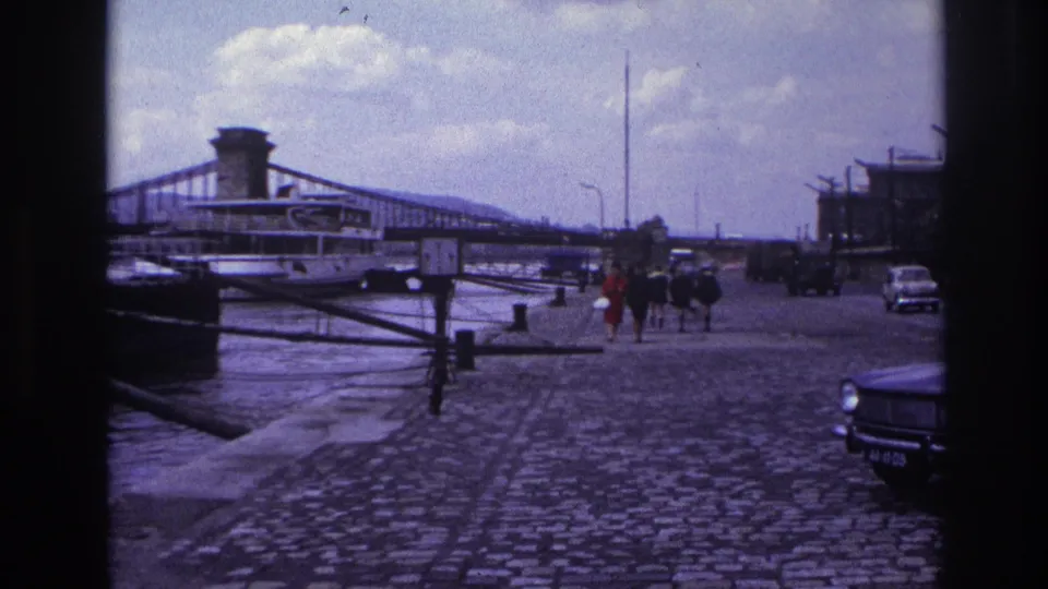 1967 France home-movie frame showing pedestrians on a cobblestone quay beside water, with a suspension bridge, riverboat, and cars visible.