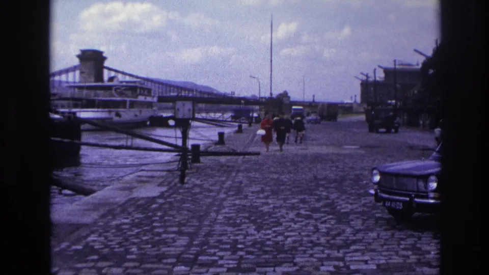 1967 France frame of people walking near the water on a cobblestone quay, with period cars, a riverboat, and a suspension bridge behind them.