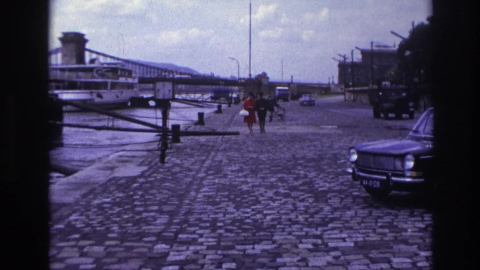A 1967 France home-movie view of a couple or pair walking along a riverside cobblestone promenade, with traffic and a suspension bridge in view.