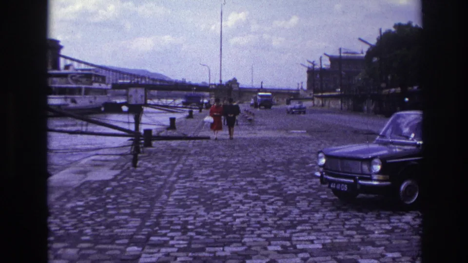 Late-1960s France home-movie frame showing two pedestrians on a cobblestone waterfront road, with a dark car in the foreground and bridge behind.