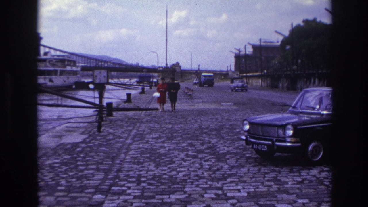 1967 France home-movie frame showing a couple walking hand-in-hand on a cobblestone riverside promenade beside a parked black sedan.
