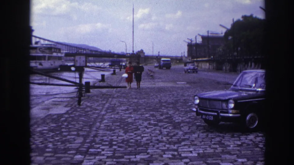 1967 France home-movie frame showing a couple walking hand-in-hand on a cobblestone riverside promenade beside a parked black sedan.