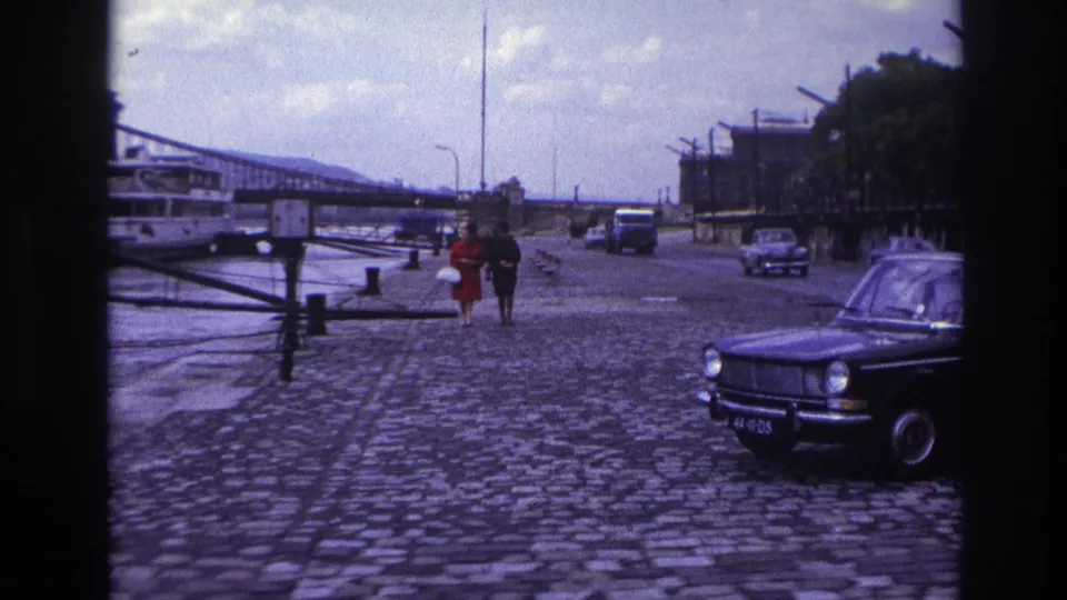 1967 France frame with a couple walking toward the camera on a cobblestone riverfront near a classic parked car and moored boat.