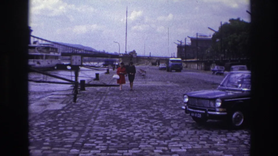 1967 France home-movie still of a couple walking along the cobblestone riverfront with traffic, a boat, and bridge behind them.