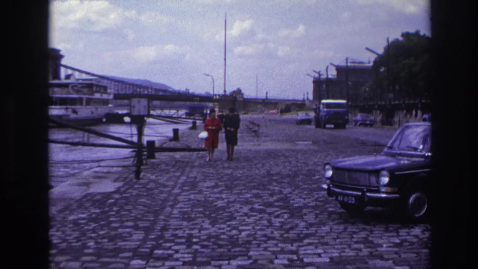 1967 France frame showing a couple on a riverside cobblestone promenade, with a dark sedan, riverboat, bridge, and traffic visible.