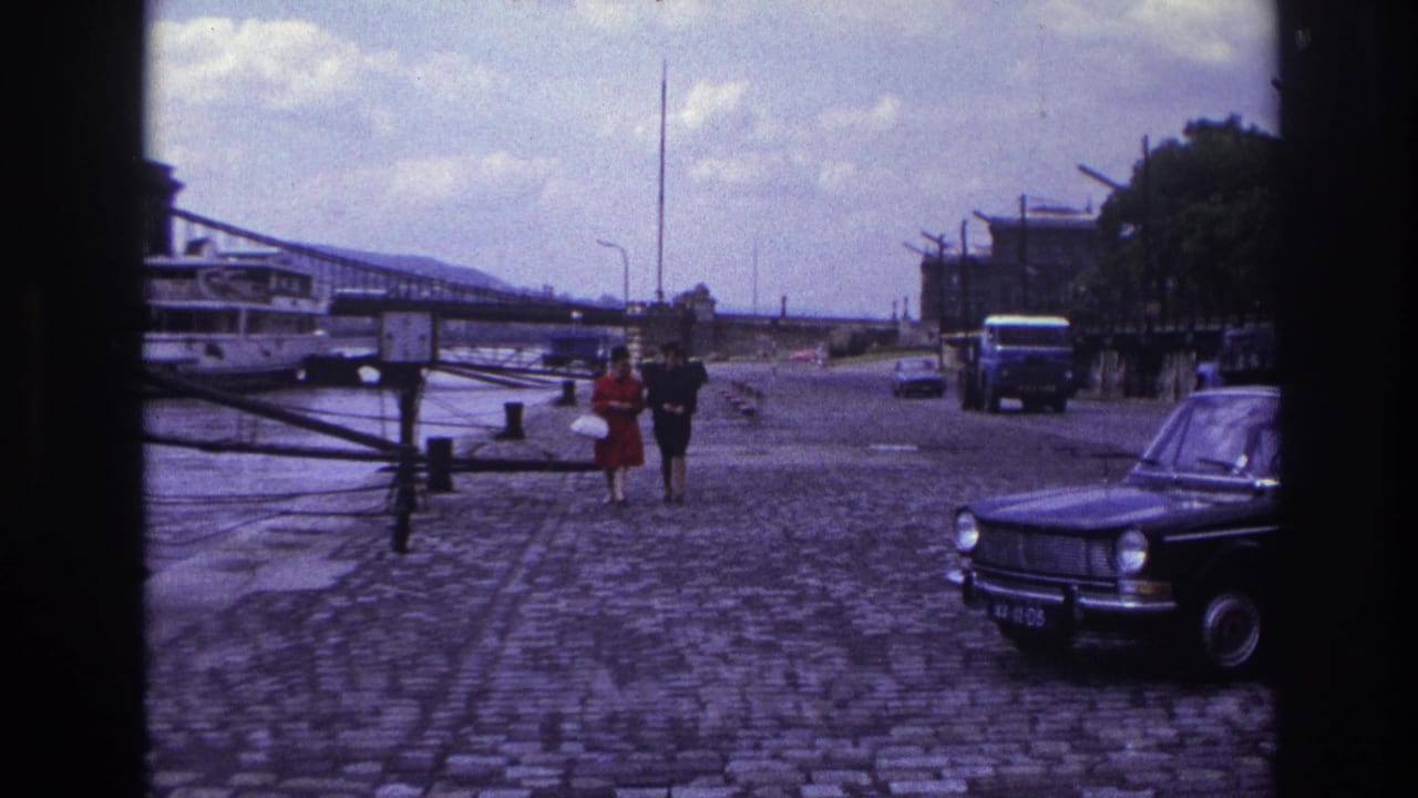 1967 France home-movie frame of two women walking toward the camera on a cobblestone riverside promenade with cars and a bridge nearby.