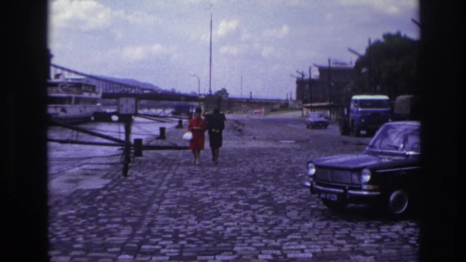 1967 France frame showing two women walking beside a riverfront roadway, with a truck, parked sedan and bridge in the background.