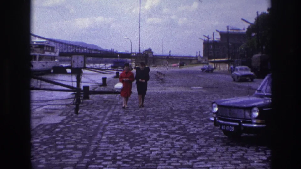 1967 France home-movie view of two women walking on a cobblestone quay, with parked cars, a bridge and riverside structures visible.
