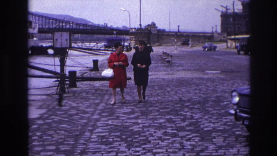 1967 France frame with two women near the camera, one in a red dress with a white handbag and one in black, on a cobblestone riverfront.