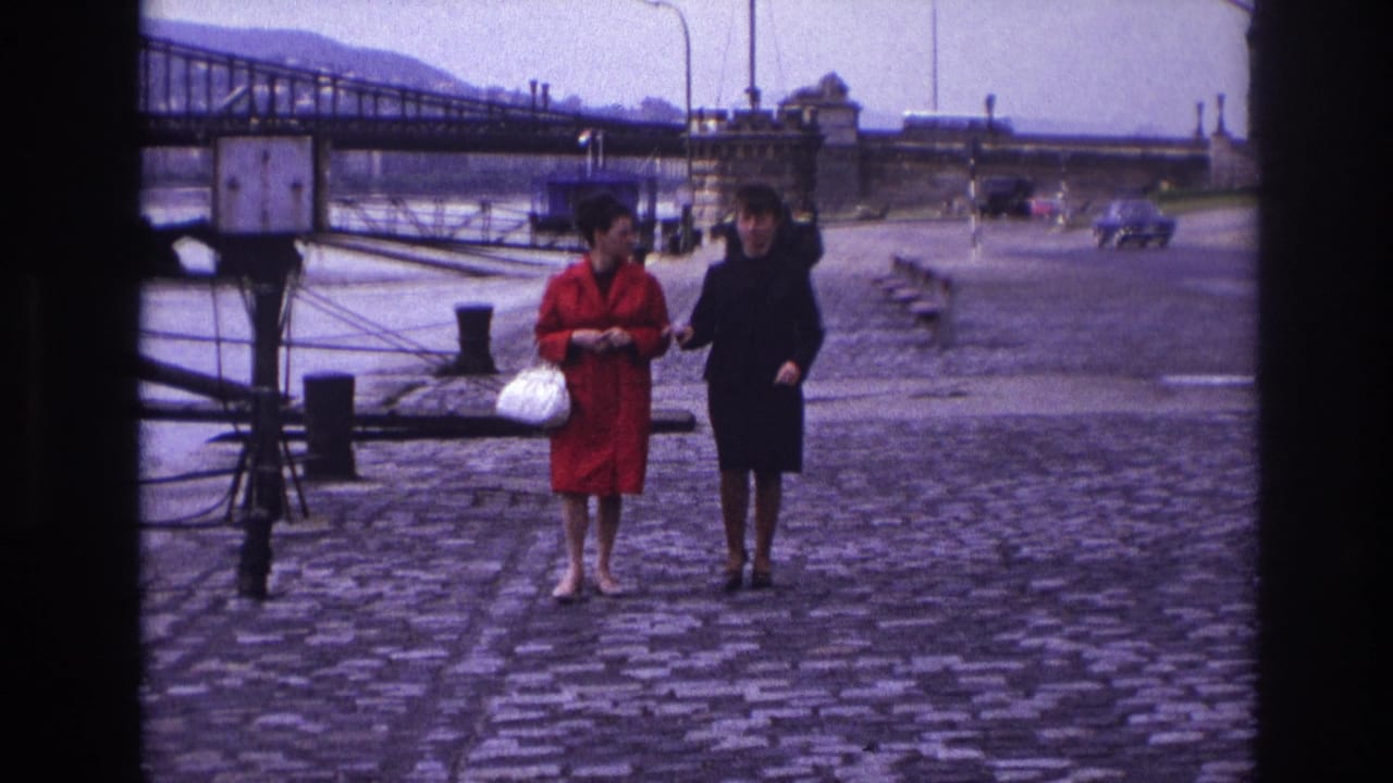 In France in 1967, two women walk arm-in-arm on cobblestones by a river, with a bridge and distant vehicles behind them.