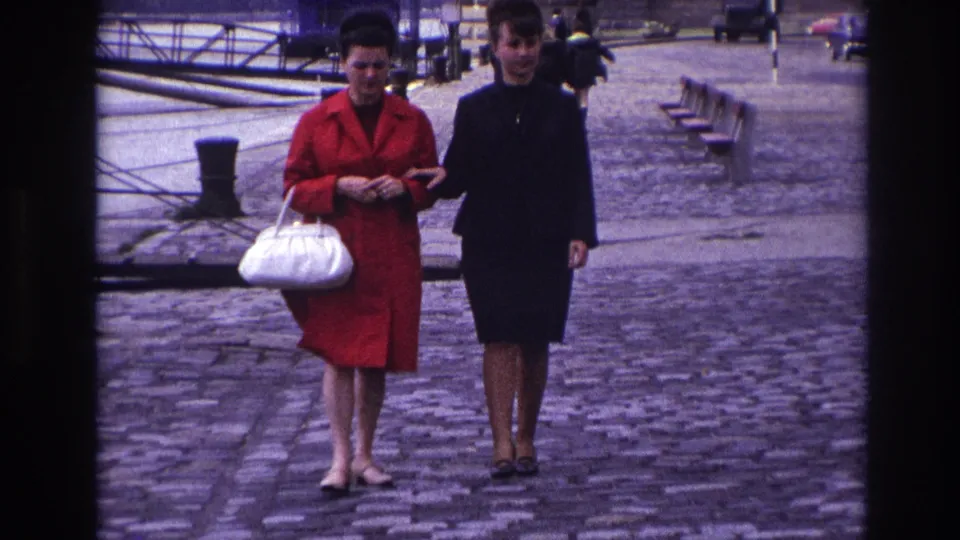 The pair faces the camera on a French riverside promenade, one in a red coat with a white handbag and one in a dark outfit.