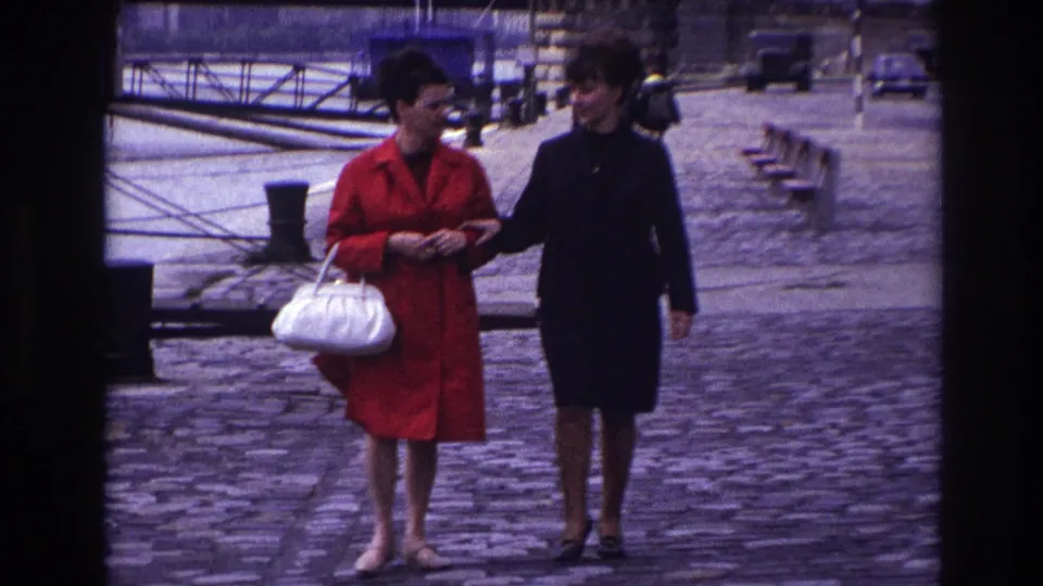 The women continue arm-in-arm in a candid 1967 France street moment, framed by cobblestones, bridge railings, and soft 8mm color.