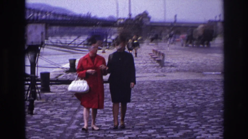The pair walks along the French riverside cobblestones in 1967, with bridgework, bollards, and distant period vehicles in view.