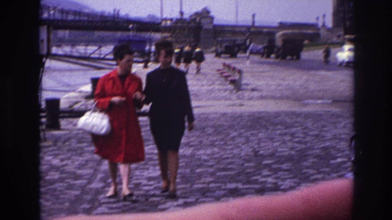 In 1967 France, two women, one in a red coat with a white handbag, walk arm-in-arm on a cobblestone waterfront street.