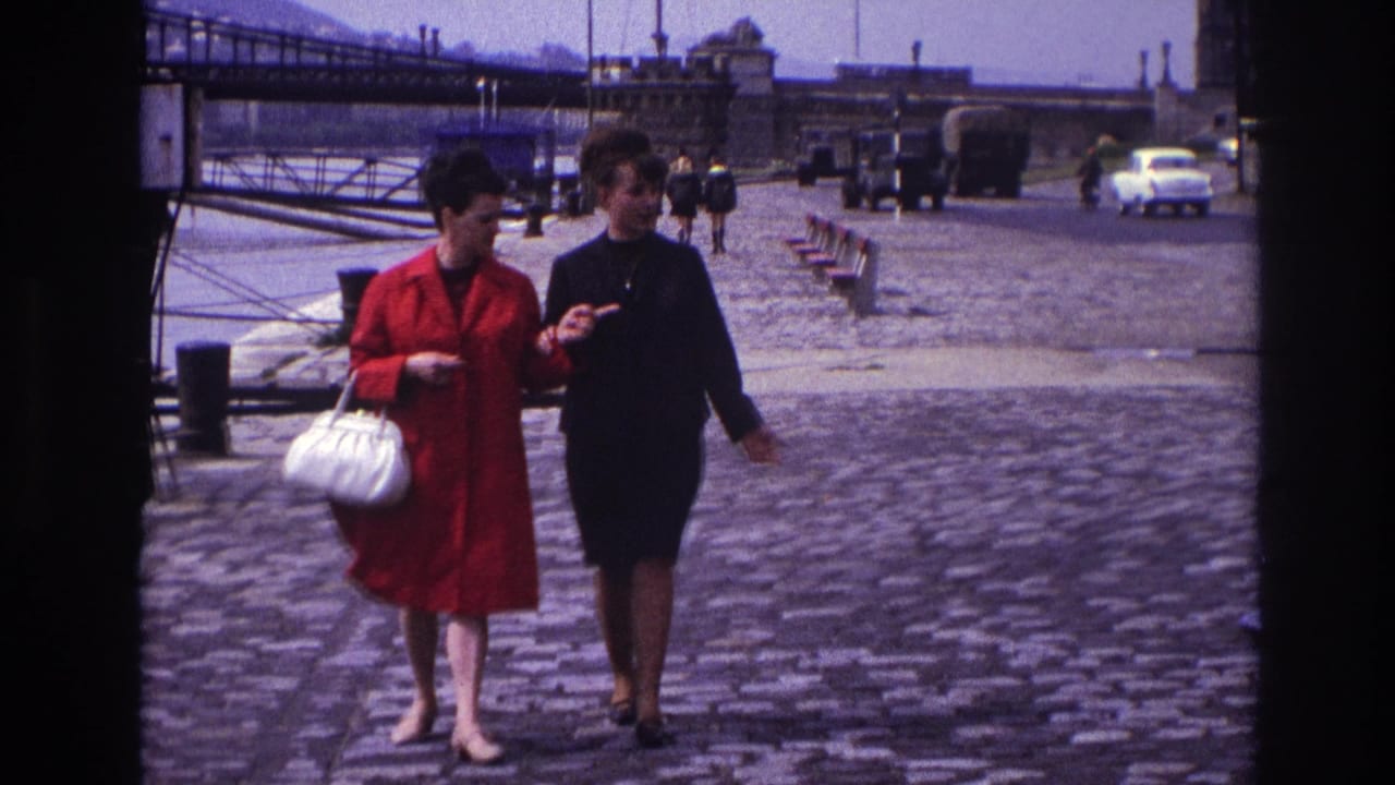 Two women link arms while walking on cobblestones with a bridge and cars behind them.