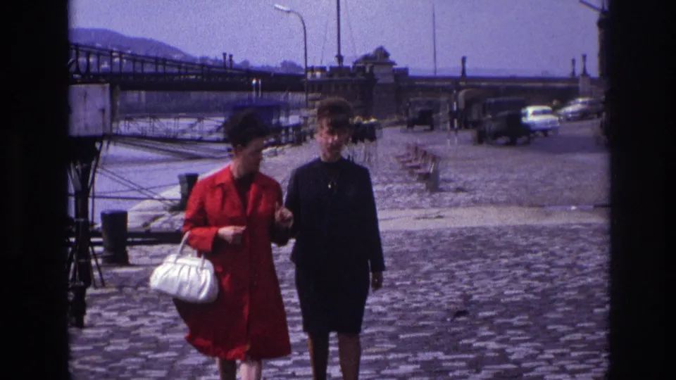 The pair continues walking together on a French waterfront street, framed by cobblestones, a bridge, pedestrians, and mid-century cars.