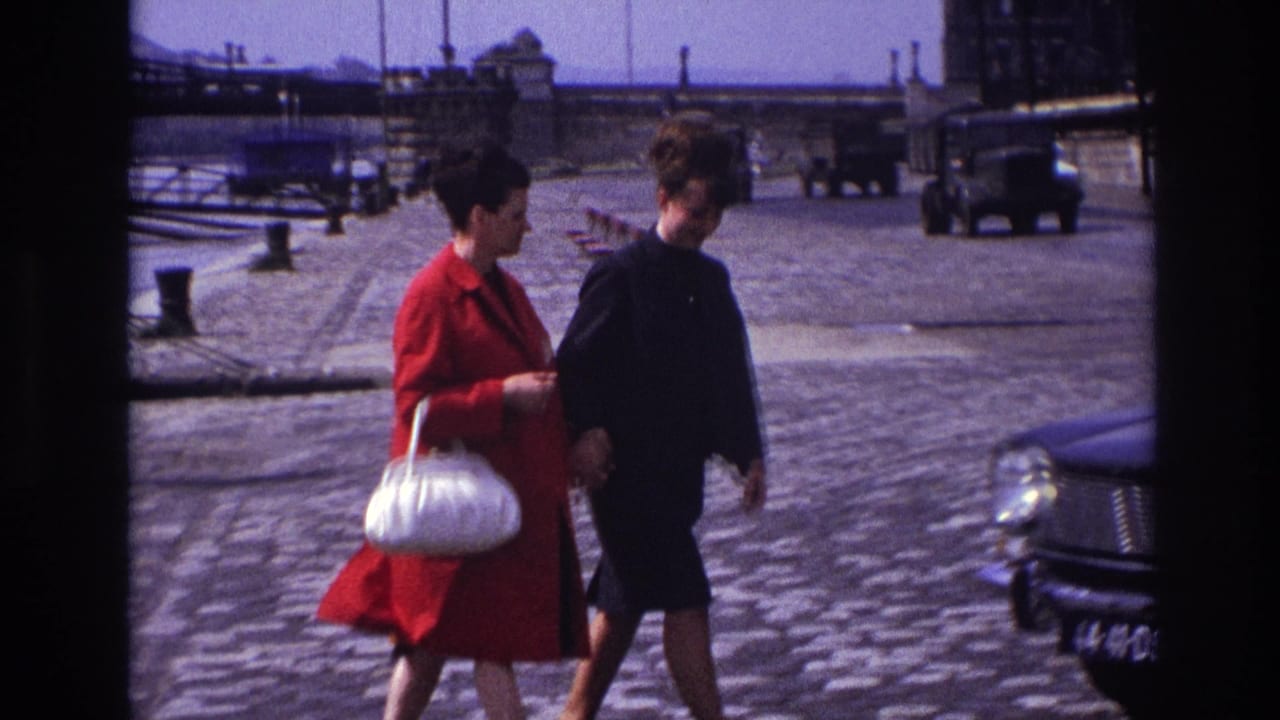 In 1967 France, two adult women walk along a cobblestone waterfront street, one wearing a bright red coat and carrying a white handbag.