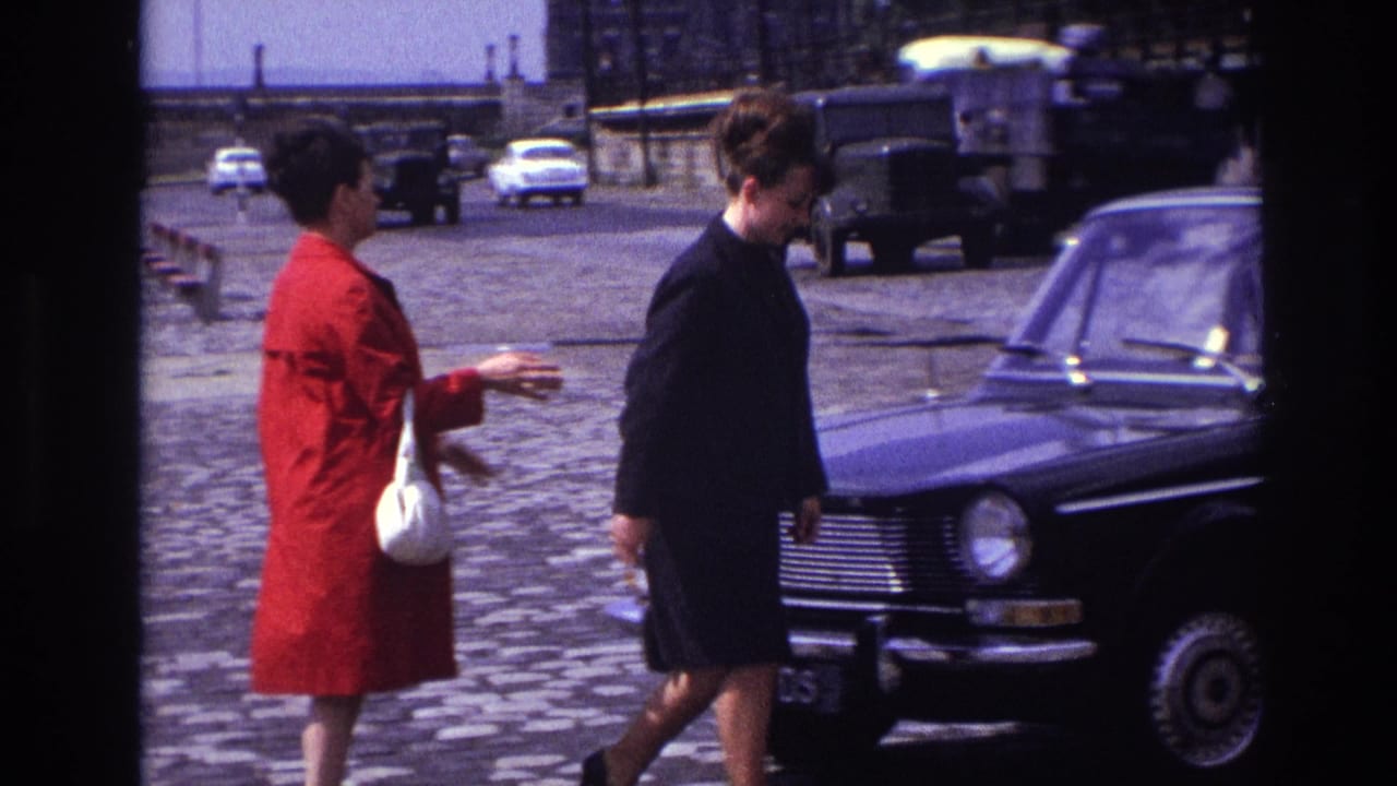 Woman in a red coat gestures beside another woman near a black sedan on a cobblestone street.