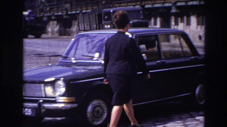 In 1967 France, a woman in a dark skirt suit walks toward the open side of a black sedan on a cobblestone waterfront road.