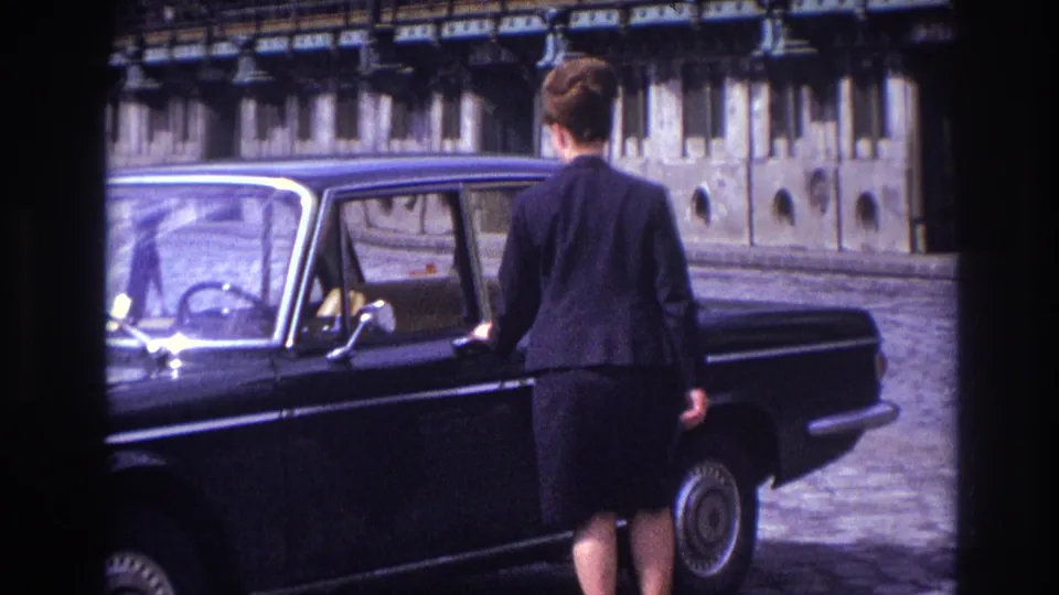In 1967 France, a woman in dark clothing stands at an open door of a black sedan beside a stone waterfront barrier.