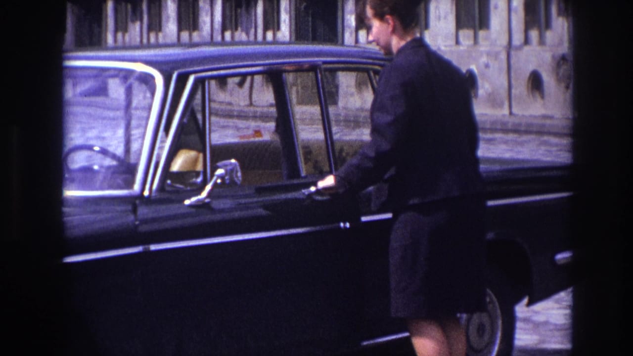 In France in 1967, a woman in dark clothing stands beside a black sedan with a stone building behind her.