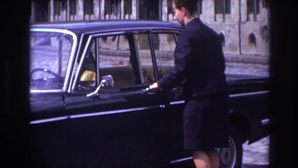 In France in 1967, a woman in dark clothing stands beside a black sedan with a stone building behind her.
